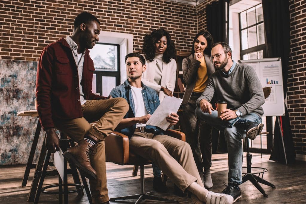 Imaginea înfățișează un grup divers de cinci profesioniști angajați într-o discuție de grup într-un birou cu ziduri din cărămidă, sugerând un mediu de lucru modern și urban. Bărbatul din stânga, îmbrăcat într-o jachetă roșie și ținând un document, stă în picioare și pare să își împărtășească opinia cu colegii, exemplificând asertivitatea. Bărbatul așezat în centru, care are în mână același document, privește în sus spre el cu o expresie de interes și considerație, indicând o comunicare eficientă. Cei trei colegi din fundal, două femei și un bărbat, privesc și ascultă activ. Această scenă sugerează colaborare, schimb de idei și respect reciproc într-un cadru de lucru dinamic și asertiv.