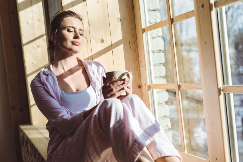 Imaginea prezintă o femeie tânără care se bucură de un moment de liniște într-un spațiu luminos, lângă o fereastră mare. Ea ține în mână o cană, probabil cu ceai sau cafea, și închide ochii, savurând căldura soarelui care pătrunde în cameră. Femeia este îmbrăcată lejer, într-o cămașă de culoare deschisă și pantaloni confortabili, semn al unei dimineți relaxate acasă. Această scenă evocă un sentiment de bunăstare și relaxare, reflectând importanța momentelor de calm și a încărcării personale în cultivarea unei vieți echilibrate și împlinite. Bunăstarea, Atmosfera plăcută și lumina naturală abudentă subliniază conexiunea dintre confortul fizic și cel mental.