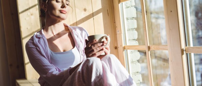 Imaginea prezintă o femeie tânără care se bucură de un moment de liniște într-un spațiu luminos, lângă o fereastră mare. Ea ține în mână o cană, probabil cu ceai sau cafea, și închide ochii, savurând căldura soarelui care pătrunde în cameră. Femeia este îmbrăcată lejer, într-o cămașă de culoare deschisă și pantaloni confortabili, semn al unei dimineți relaxate acasă. Această scenă evocă un sentiment de bunăstare și relaxare, reflectând importanța momentelor de calm și a încărcării personale în cultivarea unei vieți echilibrate și împlinite. Bunăstarea, Atmosfera plăcută și lumina naturală abudentă subliniază conexiunea dintre confortul fizic și cel mental.