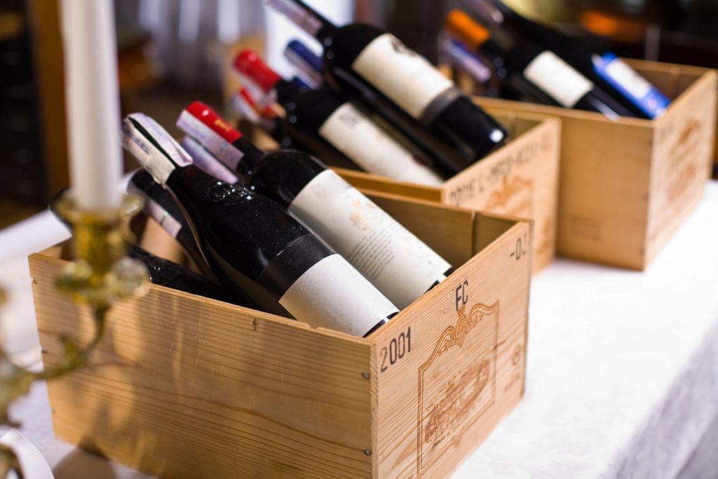 Imaginea prezintă mai multe lăzi din lemn pline cu sticle de vin, etichetate divers, sugerează diversitatea și bogăția culturii vinului. Lăzile par a fi așezate pe o masă cu o față de masă albă, creând un fundal elegant și rustic pentru prezentarea vinurilor. Această imagine poate ilustra o colecție de vinuri din diferite regiuni sau ani, reflectând pasiunea și dedicația pentru vinificație și degustarea vinului.