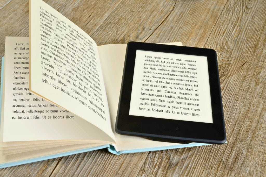 Imaginea arată un eBook reader așezat pe o carte deschisă, ambele plasate pe o suprafață de lemn. eBook reader-ul afișează text pe ecranul său, imitând aspectul paginii tipărite din carte. Această imagine subliniază tranziția de la cititul tradițional pe hârtie la cititul digital, evidențiind avantajele și versatilitatea dispozitivelor eBook readers.