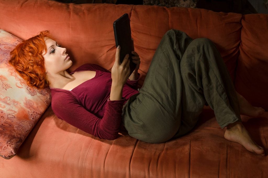 O femeie citește un eBook reader în timp ce stă confortabil pe o canapea portocalie. Ea este relaxată, bucurându-se de avantajele cititului digital oferite de ebook readers. Imaginea evidențiază portabilitatea și confortul pe care aceste dispozitive le aduc, permițând cititul în orice poziție și în diverse medii. Perfectă pentru a ilustra articolul despre beneficiile și funcțiile eBook readerelor, imaginea captează esența experienței de citire modernă și convenabilă.