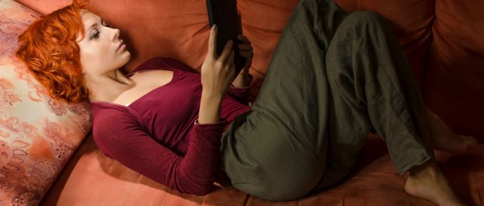 O femeie citește un eBook reader în timp ce stă confortabil pe o canapea portocalie. Ea este relaxată, bucurându-se de avantajele cititului digital oferite de ebook readers. Imaginea evidențiază portabilitatea și confortul pe care aceste dispozitive le aduc, permițând cititul în orice poziție și în diverse medii. Perfectă pentru a ilustra articolul despre beneficiile și funcțiile eBook readerelor, imaginea captează esența experienței de citire modernă și convenabilă.