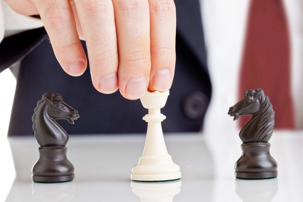 Imaginea arată o mână ce ține un pion de șah alb, pregătit să fie mutat pe tablă, cu doi cai negri de șah poziționați de o parte și de alta. Acesta poate simboliza a lua decizii strategice sau gestionarea  situații delicate, precum gestionarea conflictelor.