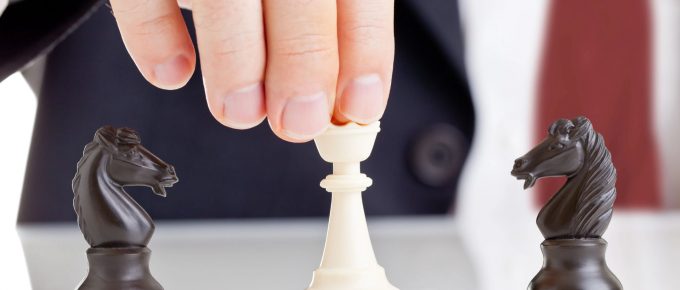 Imaginea arată o mână ce ține un pion de șah alb, pregătit să fie mutat pe tablă, cu doi cai negri de șah poziționați de o parte și de alta. Acesta poate simboliza luarea unei decizii strategice sau gestionarea unei situații delicate, precum gestionarea conflictelor.