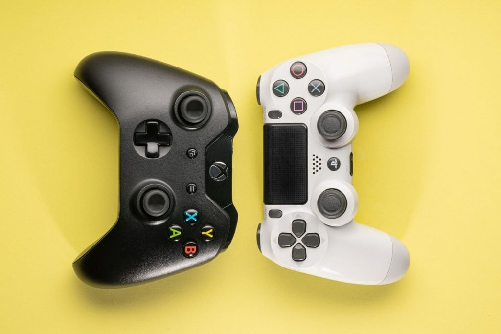 Controlere de gaming pentru console jocuri video, ilustrând un controller Xbox negru și un controller PlayStation alb pe un fundal galben, reprezentând diversitatea opțiunilor pentru jucători.