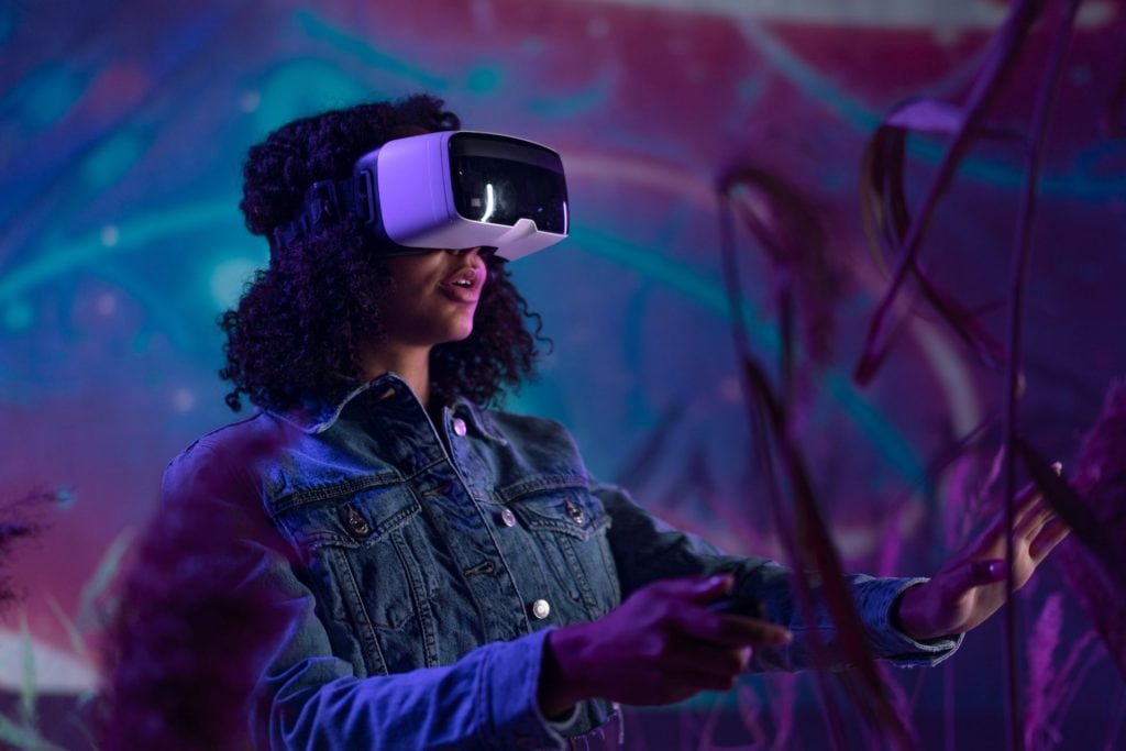 O femeie poartă ochelari VR de realitate virtuală și pare să interacționeze cu un mediu digital, mâinile ei întinse ca și cum ar atinge sau manipula obiecte invizibile. Culorile vibrante în fundal sugerează un mediu virtual plin de viață și dinamic.