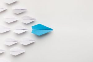 Imaginea prezintă mai multe avioane de hârtie albe dispuse într-un model regulat, cu un singur avion de hârtie albastru poziționat astfel încât să pară că conduce formația. Această configurație sugerează unicitate și inovație în leadership. Fotografia poate fi utilizată ca reprezentare vizuală a conceptului de a ieși în evidență într-un mediu de afaceri și cu abilitatea de a conduce și a inspira inovație într-un grup. Această imagine este adecvată pentru a ilustra importanța unui leadership care ghidează și influențează direcția echipei sale într-o manieră distinctă.