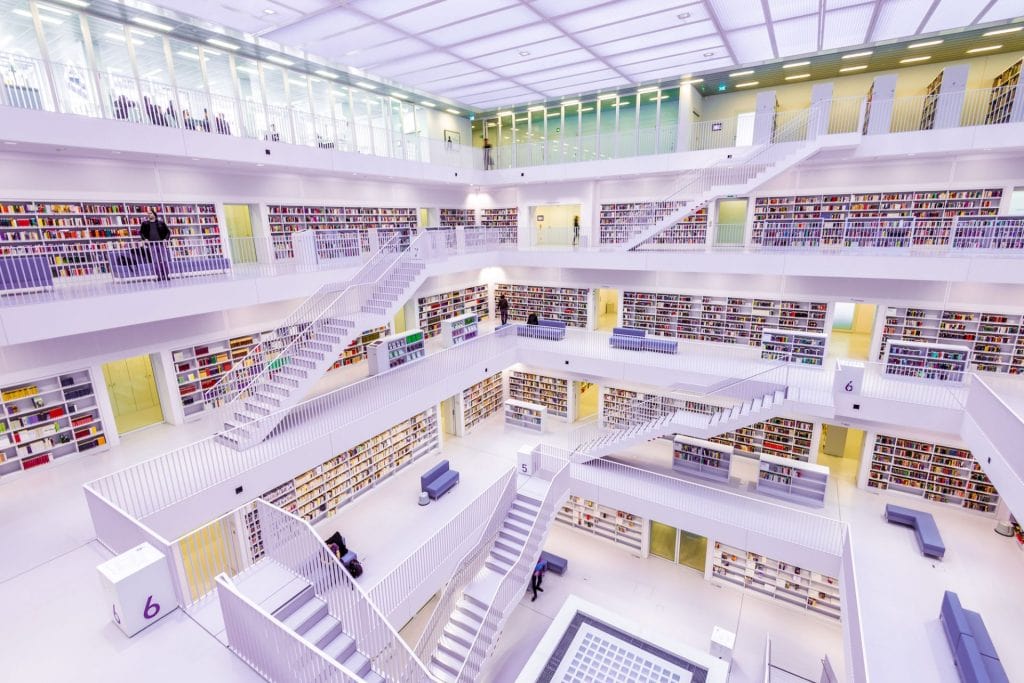 Bibliotecă modernă, spațioasă, cu mai multe etaje, rafturi albe pline de cărți și scări care le conectează. În spațiul deschis sunt câteva persoane care citesc sau se deplasează între rafturi.
