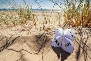 Un origami în formă de ghicitoare din hârtie plasat pe nisipul unei plaje, având inscripționate cuvintele 'WORK', 'LIFE' și 'BALANCE' scrise pe ele, sugerează echilibrul dintre viața profesională și cea personală. În fundal, se observă dune de nisip cu iarbă verde și un cer albastru.