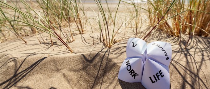 Un origami în formă de ghicitoare din hârtie plasat pe nisipul unei plaje, având inscripționate cuvintele 'WORK', 'LIFE' și 'BALANCE' scrise pe ele, sugerează echilibrul dintre viața profesională și cea personală. În fundal, se observă dune de nisip cu iarbă verde și un cer albastru.
